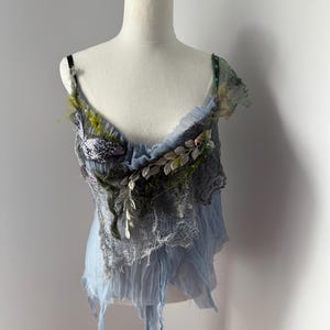 May include: A light blue, layered top with decorative embellishments. The top features a draped design with a ruffled neckline and delicate straps. Embellishments include silver leaves, green moss, and a textured, gray fabric.
