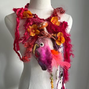 May include: A decorative fiber art neckpiece featuring a needle-felted pink parrot head with a yellow beak and white eye. The piece is adorned with vibrant magenta yarn, textured red fibers, orange fabric flowers, and a bright pink feather accent.