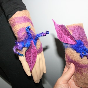 May include: A pair of felted wool wrist warmers with purple and blue accents. The warmers are decorated with felted leaves and flowers.