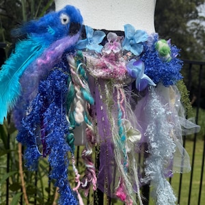 Pode incluir: Uma saia de fada artesanal com um detalhe de pássaro azul. A saia apresenta uma variedade de texturas, incluindo penas, fios e tecido em tons de azul, roxo e verde. Enfeitada com flores e elementos decorativos.