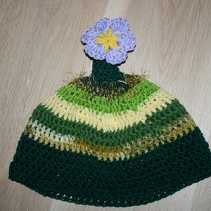 May include: A green, yellow, and brown crocheted hat with a purple flower on top. The hat is striped and has a fuzzy trim.