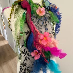 Pode incluir: Uma vibrante peça de arte têxtil feita à mão com um design central de penas rosa e azul, adornada com flores rosa, folhagem verde e uma base de renda preta. A peça é exibida em um manequim, mostrando seus detalhes intrincados e composição colorida.