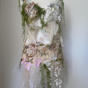 May include: A whimsical, handmade dress on a mannequin, adorned with floral embellishments and intricate lace. The bodice features a mix of sequins, beads, and faux greenery. Soft pink and white flowers are scattered across the dress, with a flowing, layered skirt.