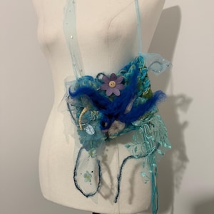 Op de afbeelding: Een handgemaakte schoudertas in blauwe en turquoise tinten met een grillig ontwerp. De tas heeft verschillende texturen, waaronder stof en garen, en een paarse bloem met een knoop in het midden. Een dunne blauwe band met pailletten maakt het ontwerp af.
