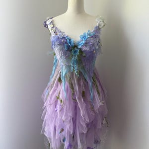 Puede incluir: Un vestido de hada lavanda con una falda de tul en capas y un corpiño adornado con detalles florales azules y morados. El vestido presenta encaje delicado y detalles de tela fluida, creando un aspecto etéreo. El vestido se exhibe en un maniquí.