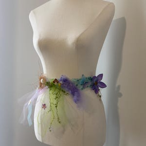 May include: A whimsical fairy-themed belt, displayed on a mannequin. The belt features a tulle skirt in shades of white, green, and lavender, adorned with faux flowers, moss, and a mushroom. The belt is embellished with purple and blue accents.