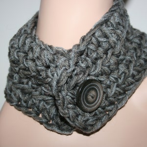 Puede incluir: Un calentador de cuello de ganchillo gris con un cierre de botón grande y oscuro. El diseño de punto grueso presenta diferentes tonos de hilo gris. Este accesorio hecho a mano es un accesorio acogedor.