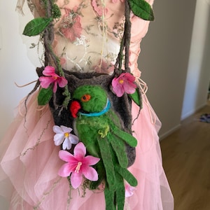 Op de afbeelding: Een handgemaakte vilten tas met een groene papegaai met een rode snavel en een blauwe kraag. De tas is versierd met roze bloemen, groene bladeren en bruine wijnranken. De tas wordt opgehangen door twee bandjes.