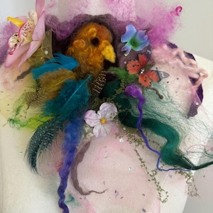 May include: A whimsical, handcrafted neckpiece featuring a felted bird with orange and yellow hues, surrounded by colorful feathers, flowers, and tulle. The design incorporates shades of pink, purple, blue, and green, creating a vibrant and artistic accessory.