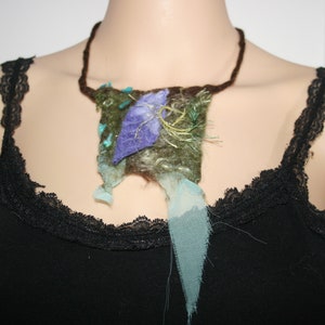 Può includere: Una collana di feltro marrone e verde con un ciondolo a forma di fiore blu e verde. La collana è su un manichino che indossa una canotta nera con bordino in pizzo.