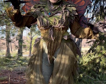 May include: A person in a forest-themed costume with intricate makeup and headpiece. The costume features green and brown fabrics, with a large, ornate wing-like structure. The person is posing in a natural outdoor setting.