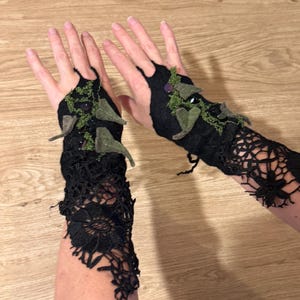 Può includere: Guanti neri senza dita con pizzo intricato e accenti di foglie di feltro. I guanti presentano una combinazione di colori verde scuro e viola, con un design in pizzo floreale che si estende sull'avambraccio. Decorati con foglie di feltro verdi e piccole perle viola.