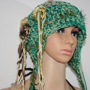 May include: A green, brown, and white crocheted hat with long strands of yarn hanging down. The hat has a flower design on the side.