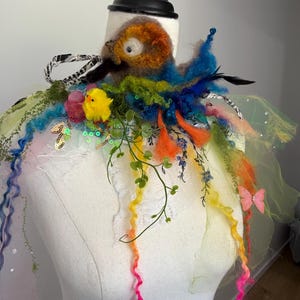 May include: A whimsical, colorful neckpiece with a felted bird design, featuring orange, blue, and green accents. The piece includes a yellow chick, pink butterflies, and trailing curly ribbons in rainbow hues. A black and white ribbon adds a contrasting detail.