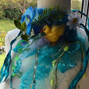 Pode incluir: Um colar artesanal e caprichoso com um patinho amarelo feltrado aninhado entre lã azul e turquesa. Adornado com flores azuis, folhas verdes e uma margarida branca, a peça é acentuada com fitas verde-azuis e delicados fios de ouro e prata.