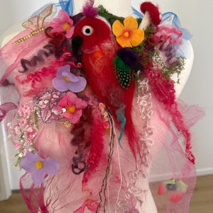 May include: A decorative textile accessory featuring a needle-felted red parrot, vibrant pink and purple fabric layers, artificial flowers, green moss accents, and a sheer blue ribbon. The piece includes sequins, curly fibers, and a small butterfly.