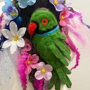 Könnte beinhalten: Ein dekorativer Kragen mit einem grünen Papagei aus Filz mit rotem Schnabel und orangefarbenem Auge. Der Kragen ist mit bunten Stoffblumen in Rosa, Blau und Weiß verziert und wird auf einer Schaufensterpuppe präsentiert.