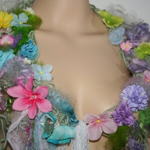 May include: A colourful floral necklace made of yarn, fabric, and faux flowers. The necklace features a variety of colours, including pink, blue, green, and purple. The flowers are arranged in a cascading pattern, creating a whimsical and romantic look.