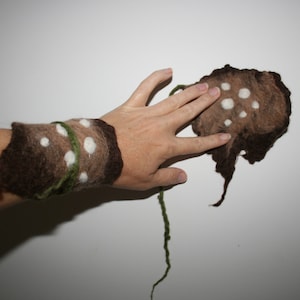 May include: A brown and tan felted wrist cuff with white polka dots and a green felt accent. The cuff is shown on a person's wrist.