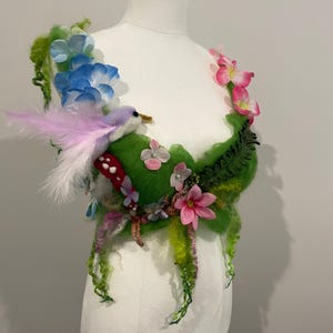 Puede incluir: Un corpiño de fieltro verde adornado con un pájaro, flores y vegetación. El corpiño está decorado con flores azules, rosas y blancas, así como hojas y enredaderas verdes. Un pequeño pájaro con plumas rosas está posado en un hongo rojo.