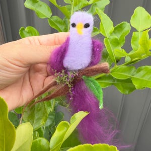 Peut inclure: Un oiseau en feutre violet avec un bec jaune et des yeux noirs perché sur une branche brune avec des feuilles vertes. L'oiseau a une longue queue violette.