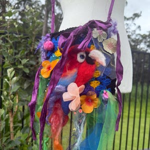 Op de afbeelding: Een kleurrijke, handgemaakte schoudertas met een vogelontwerp. De tas heeft een rode, blauwe en gele vogel, versierd met stoffen bloemen en linten. De tas heeft een zwarte basis en is versierd met groene, blauwe en oranje tule.