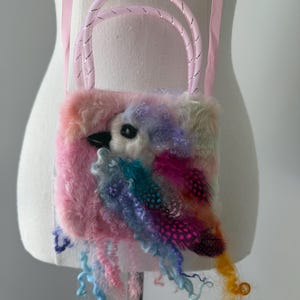 Op de afbeelding: Een handgemaakte tas in de vorm van een vogel, getoond op een mannequin. De tas heeft een pluizig, veelkleurig ontwerp met roze, blauwe en paarse tinten, geaccentueerd met veren en krullend garen. Het heeft een roze handvat en een bijpassende riem.
