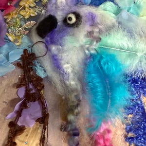 Pode incluir: Close-up de uma obra de arte têxtil com uma criatura caprichosa com um olho preto e um corpo fofo e texturizado em tons de roxo, azul e branco. A obra de arte incorpora penas, flores de tecido e lantejoulas, criando uma composição vibrante.