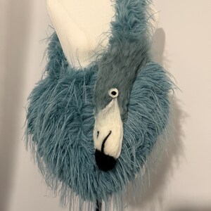 Op de afbeelding: Een unieke, blauwe gevederde handtas in de vorm van een flamingo. De tas heeft een witte en zwarte vilten snavel en oog, met een pluizig, getextureerd lichaam. De tas wordt op een mannequin gepresenteerd en toont het unieke ontwerp.
