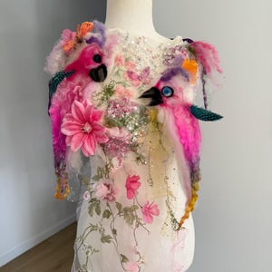 Op de afbeelding: Een wit mouwloos kledingstuk met twee naaldgevilte roze en paarse vogels met donkere snavels en blauwe ogen. Het stuk is versierd met roze stoffen bloemen, lovertjes, lagen van doorschijnende stof en kleine vlinderaccenten aan de voorzijde.