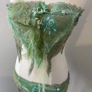 May include: A green and teal fairy costume, comprising a top and a matching band. The top is adorned with lace, embroidery, and fabric with a distressed appearance. The costume is displayed on a mannequin.