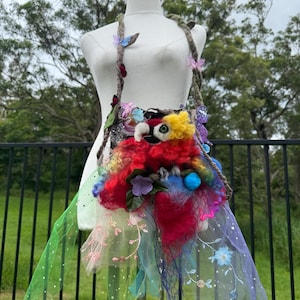 Op de afbeelding: Een grillige, handgemaakte schoudertas met een papegaai-ontwerp. De tas heeft levendige kleuren, waaronder rood, geel, blauw en groen, met bloemen- en vlinderversieringen. De riem heeft een gedraaid touwontwerp en de tas heeft een tule rok.