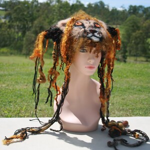 Pode incluir: Um adorno de cabeça de leão feito à mão com um rosto detalhado e detalhes de estilo dreadlock laranja e preto. O adorno de cabeça é exibido em uma cabeça de manequim, mostrando o intrincado design de lã feltrada. O fundo é um campo gramado.