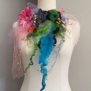 Op de afbeelding: Een grillig, handgemaakt halssieraad met een groene vogel met een gele snavel, blauwe veren en kleurrijke bloemen. Het ontwerp bevat roze, oranje en blauwe accenten, met een doorschijnende, geborduurde stof.