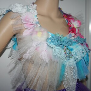 May include: A vibrant, handmade bodice with a floral design. The top combines lace, tulle, and crochet, embellished with fabric flowers in pink, blue, and white. The bodice is displayed on a mannequin, showcasing its intricate details.