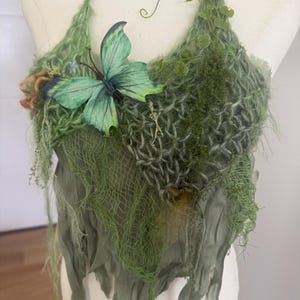 Può includere: Un top verde all'uncinetto con dettagli di farfalle e foglie. Il top presenta un design intrecciato con tessuto drappeggiato e elementi vegetali decorativi. Una grande farfalla verde è attaccata sul davanti.