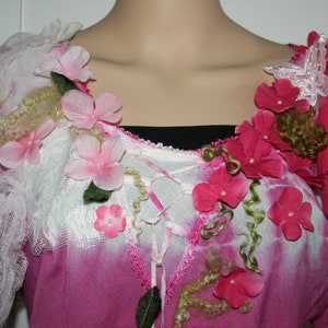 May include: A pink shirt with a white lace overlay and pink and red fabric flowers. The flowers are arranged in a cluster around the neckline and are accented with green leaves and brown curly fibres.