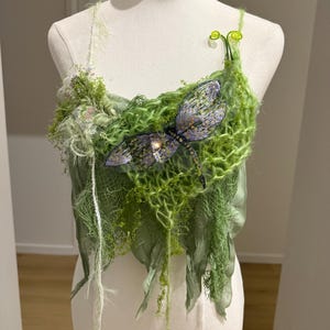 Può includere: Top verde fatto a mano con un design a libellula. Il top presenta un mix di trame, tra cui rete, foglie di tessuto e delicati abbellimenti. La libellula è finemente dettagliata con accenti viola e oro, aggiungendo un tocco di fantasia all'indumento.