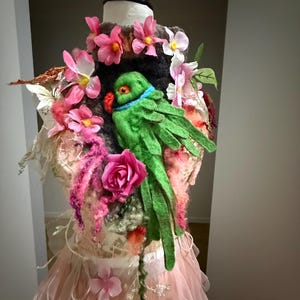 Op de afbeelding: Een decoratieve paspop versierd met een vilten groene papegaai, roze bloemen en een roze tule rok. De papegaai heeft een blauwe halsband en een rode snavel. De paspop is versierd met diverse roze en witte bloemen.