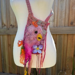 Op de afbeelding: Handgemaakte roze en paarse gehaakte crossbodytas met een vilten vogelontwerp. De vogel heeft een gele snavel en zwarte ogen, met blauwe bloemen en paddenstoelversieringen. De tas wordt getoond op een witte mannequin.