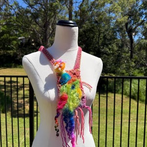 Op de afbeelding: Een kleurrijke, handgemaakte ketting op een paspop. De ketting heeft een haakontwerp met levendige tinten roze, geel, groen, blauw en paars. Het heeft een getextureerde, gefranjerde hanger en een gevlochten band.