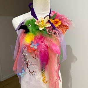 Op de afbeelding: Een decoratief draagbaar kunstobject met een gevilte groene papegaai met een zwarte snavel, gelaagd met roze en paarse tule, zijden linten, kunstbloemen, veren, pailletten en vlinderapplicaties, vastgezet met een paarse stoffen nekband.