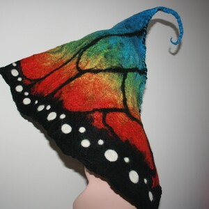 Puede incluir: Un sombrero de fieltro con forma de ala de mariposa con un patrón de arcoíris y detalles en blanco y negro. El sombrero tiene una parte superior puntiaguda y una cola larga y rizada.