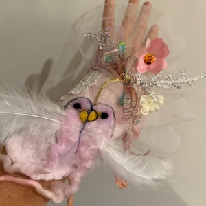 May include: A pink and white feathered creature with a yellow beak and black eyes, wearing a white tulle veil with pink flowers and silver glitter. The creature is attached to a person's arm.