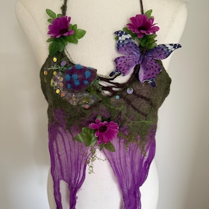 Può includere: Un top all'americana verde oliva scuro con una farfalla viola, due fiori magenta, muschio artificiale verde, cerchi in feltro blu e paillettes iridescenti. Il capo presenta un laccetto marrone al collo e un orlo in tessuto viola sfrangiato.