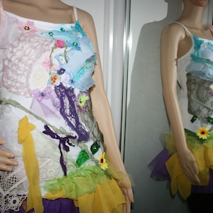 May include: A white tank top with a variety of colorful fabric scraps and flowers sewn on. The top has a yellow, green, and purple layered skirt.