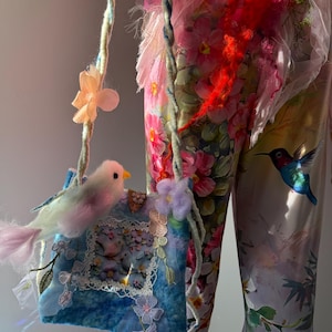 Op de afbeelding: Een grillige, handgemaakte blauwe tas met een gevilte vogel erop. De tas heeft bloemversieringen, een met kant afgezette foto en een gedraaide koordriem versierd met bloemen. De achtergrond bevat stof met bloemenpatroon.