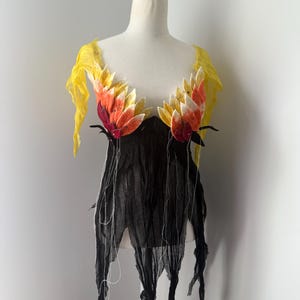 Può includere: Un capo unico, fatto a mano, esposto su un manichino. La parte superiore presenta un corpetto nero e strappato con lunghe strisce pendenti. Il busto è adornato da grandi accenti floreali colorati nelle tonalità del rosso, dell'arancione e del giallo.
