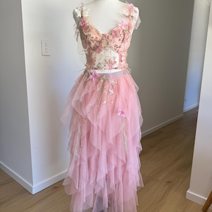 May include: A two-piece formal gown on a mannequin. The bodice is a light pink with floral and butterfly accents. The skirt is a layered, ruffled tulle in a matching pink. The dress is displayed on a black mannequin stand against a white wall.