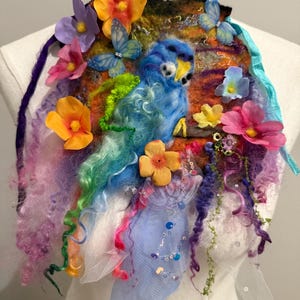 Op de afbeelding: Een kleurrijk, handgemaakt viltkunstwerk met een blauwe vogel met een gele snavel, omringd door levendige bloemen, vlinders en vloeiende, regenboogkleurige wol. Het stuk wordt op een mannequin getoond.